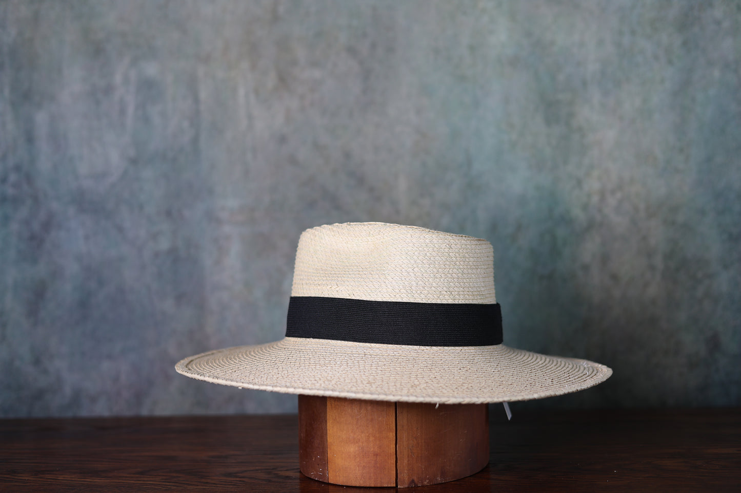 Beige straw hat with a black band on a wooden block against a textured blue background