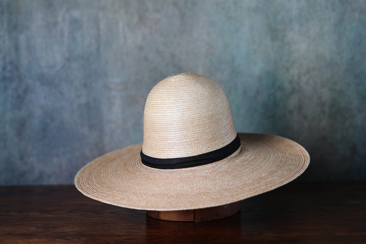 Beige straw hat (Sunbody Oak Open Crown Palm Hat 5" Brim) with a black band on a wooden stand against a textured gray background